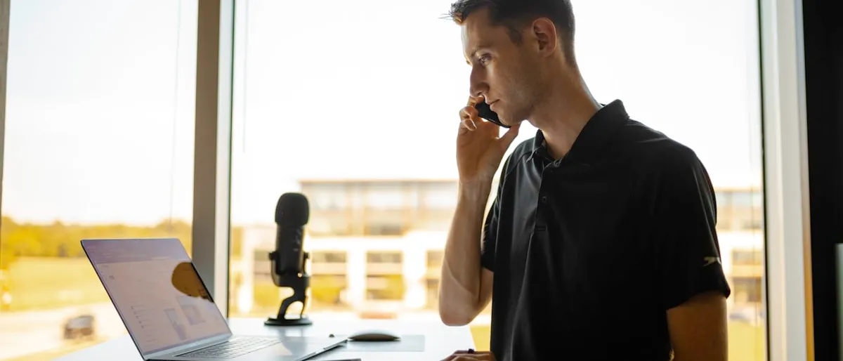 A man on a phone call while standing at his desk with a laptop open