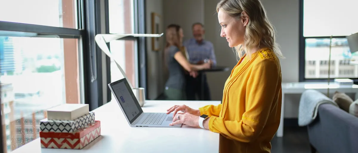 A woman typing on a laptop at a standing desk near a window